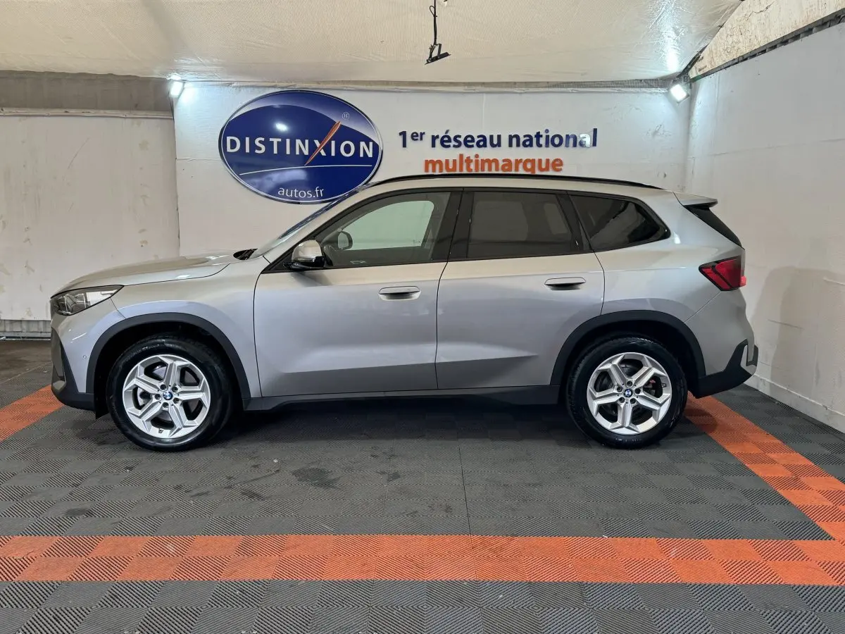 Vue de profil côté gauche d'un BMW X1 gris foncé 2024 dans un showroom avec logo Distinxion en arrière-plan.