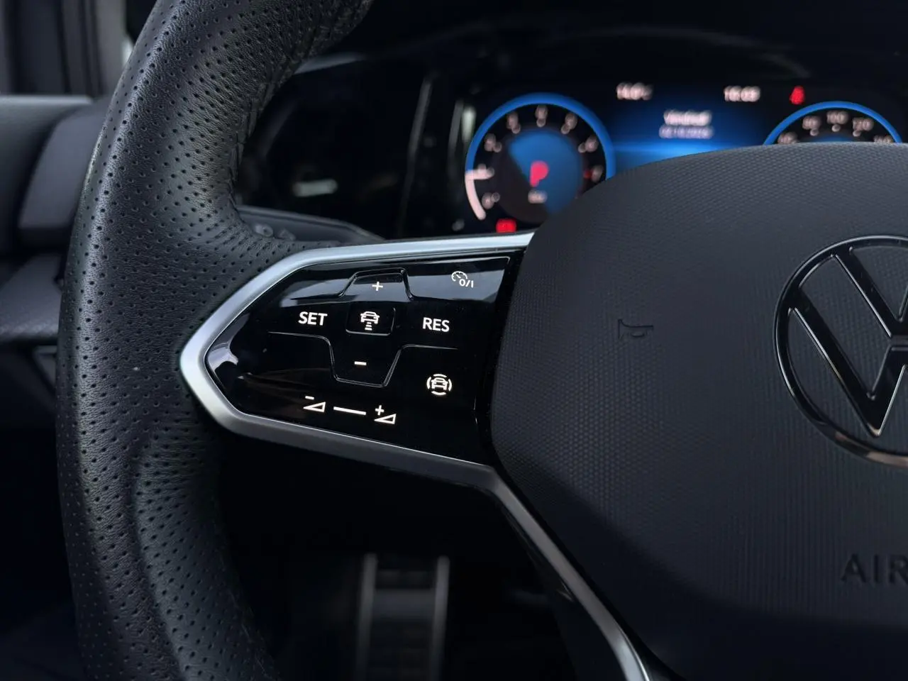 Gros plan sur le volant cuir noir perforé de la Volkswagen Golf 8 R-Line avec commandes multifonctions et tableau de bord digital bleu.