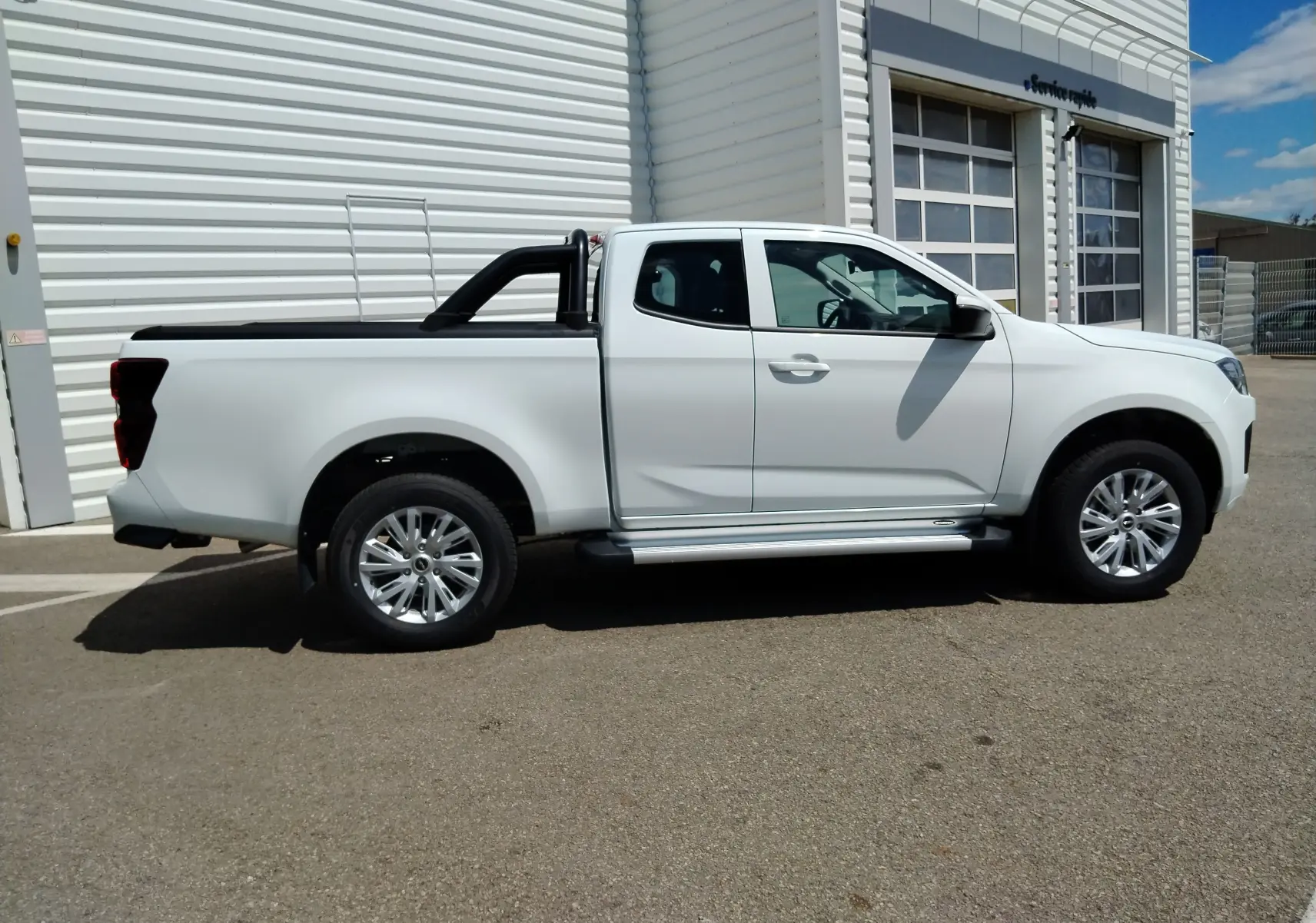 Vue de profil côté droit d'un pick-up Isuzu D-MAX blanc avec arceau noir et couvre-benne roulant, stationné devant un bâtiment industriel.