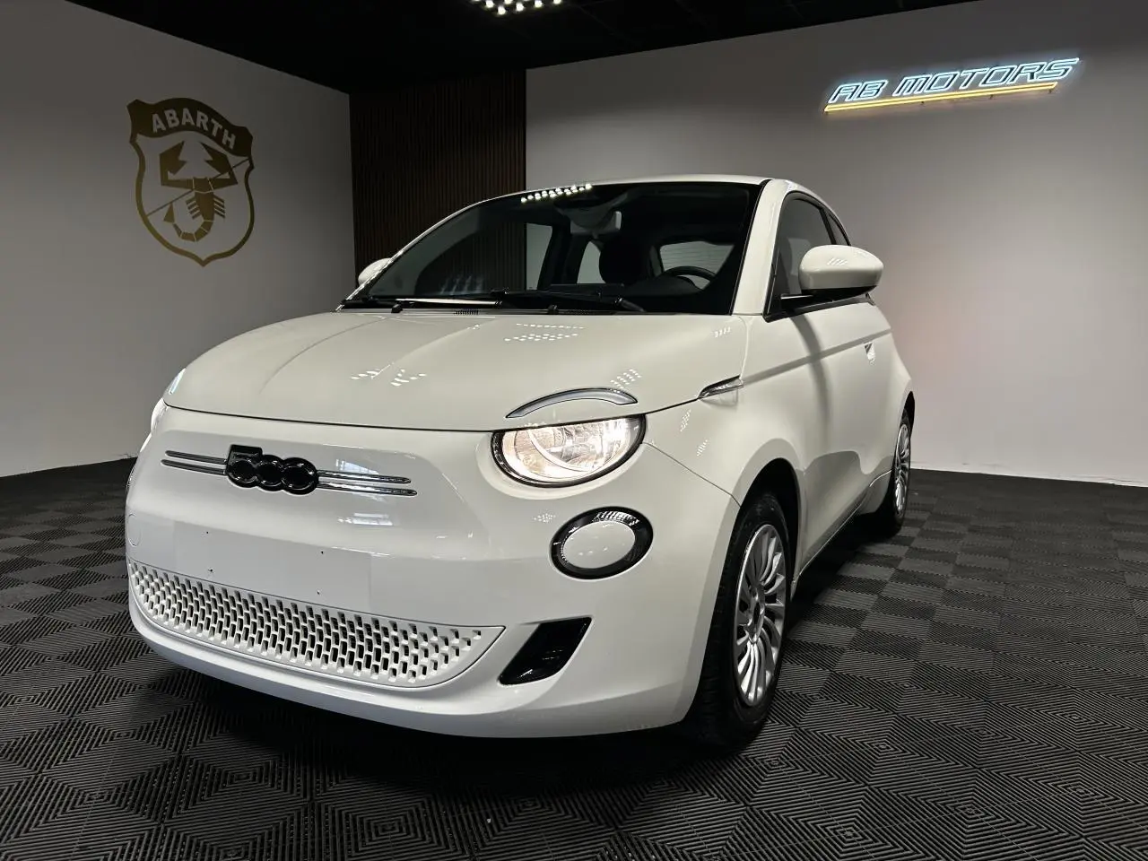 Fiat 500 électrique blanche vue 3/4 avant droit dans un showroom avec phares allumés et jantes acier.