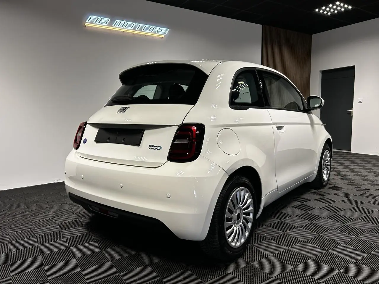 Vue 3/4 arrière droite d'une Fiat 500 électrique blanche 2024 avec jantes acier et feux arrière foncés en intérieur showroom.