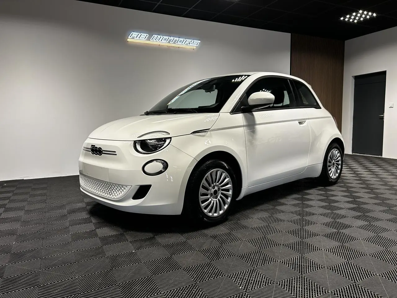 Vue 3/4 avant droite d'une FIAT 500 électrique blanche 2024 avec jantes acier et éclairage LED diurne en showroom.