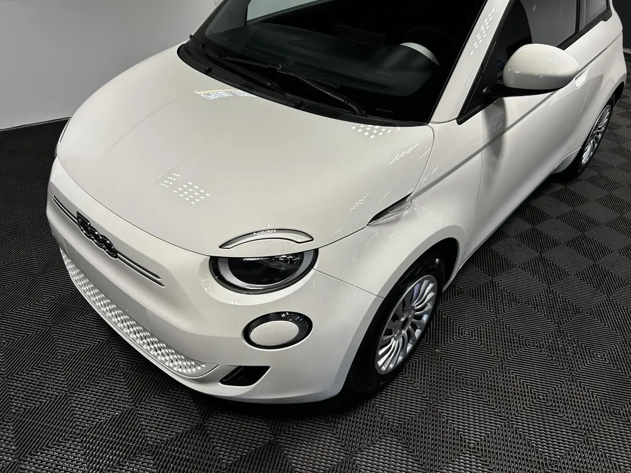 Vue 3/4 avant droit d'une FIAT 500 électrique blanche 2024 avec jantes acier et phares ronds distinctifs.