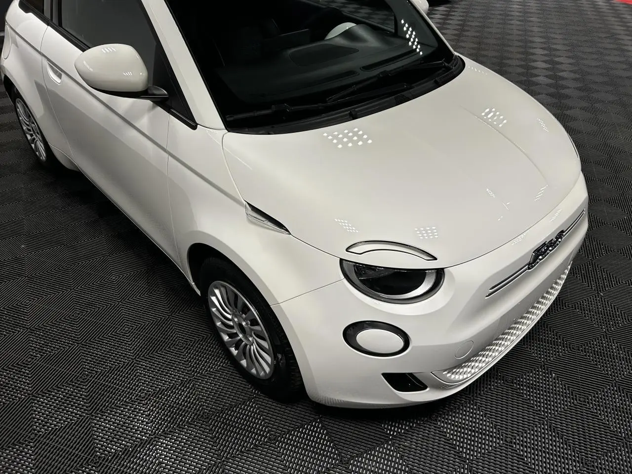 Vue 3/4 avant gauche d'une Fiat 500 électrique blanche 2024 avec jantes acier et phares ronds LED.