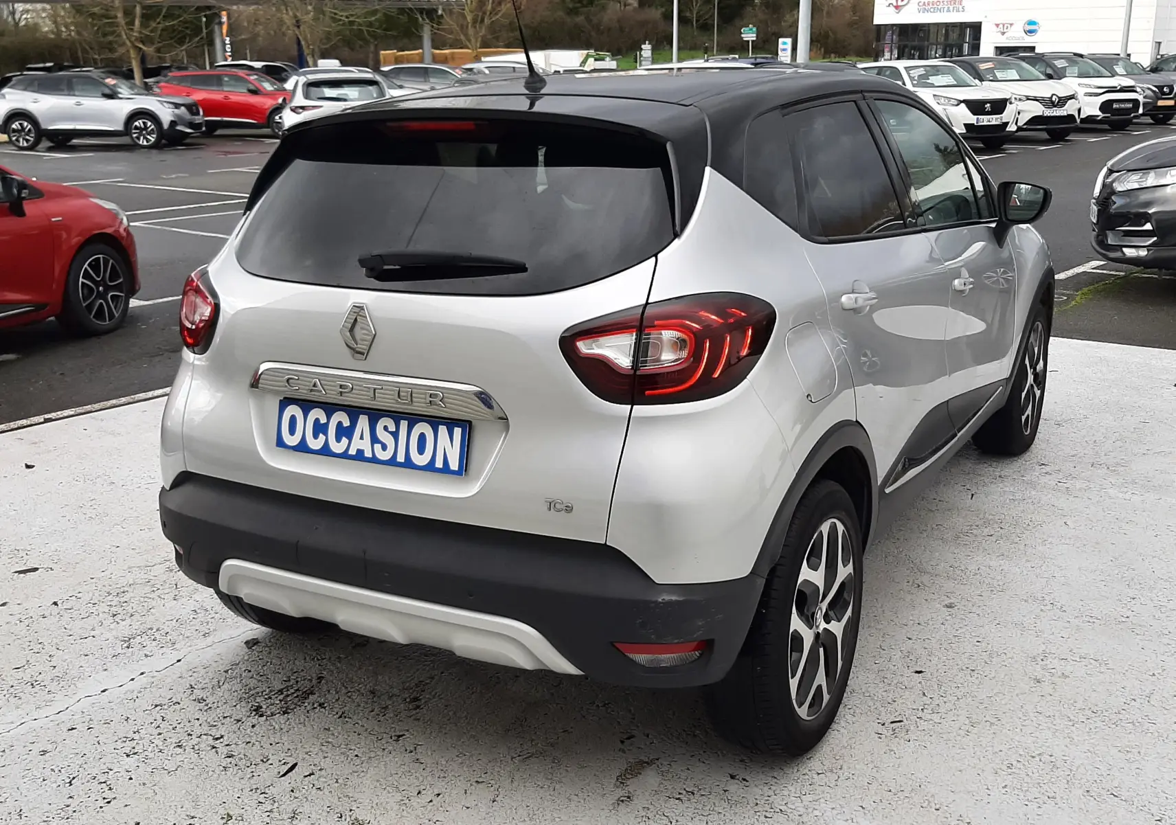 Vue 3/4 arrière droite d'un Renault Captur gris avec toit noir, feux arrière allumés, sur parking extérieur.