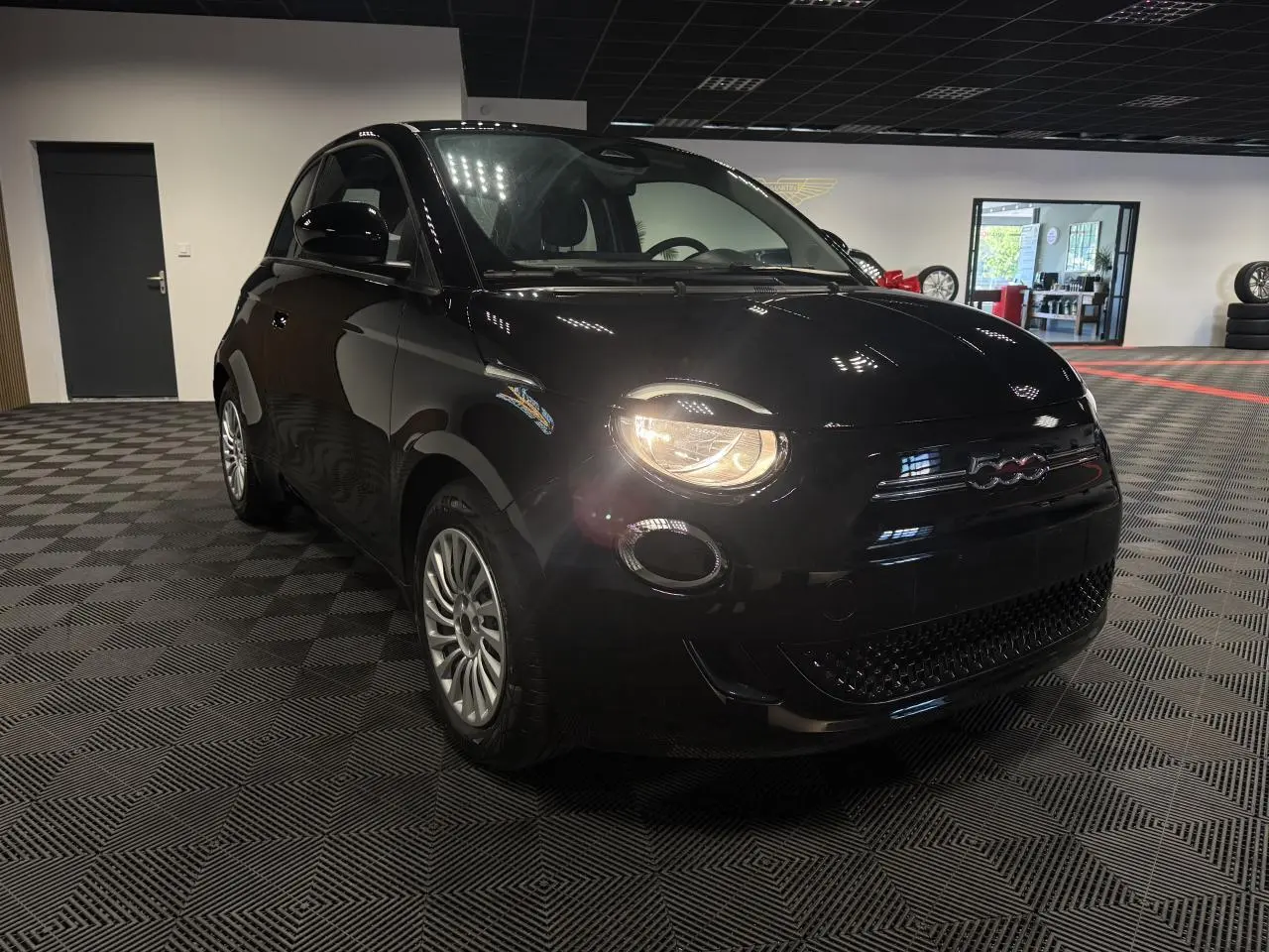 Fiat 500 électrique noire vue en 3/4 avant droit avec phares allumés dans un showroom intérieur.