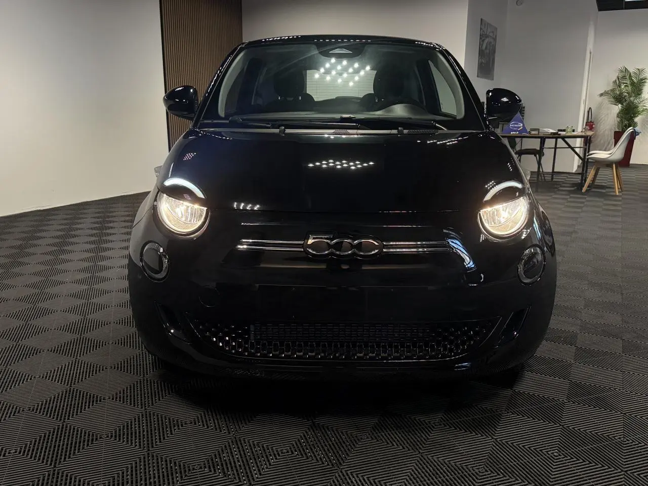 Vue de face d'une FIAT 500 électrique noire avec phares allumés dans un intérieur showroom.