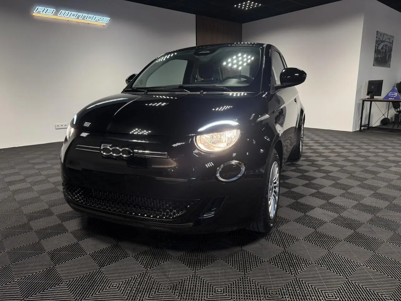 Fiat 500 électrique noire vue 3/4 avant droit avec phares allumés dans un showroom intérieur.