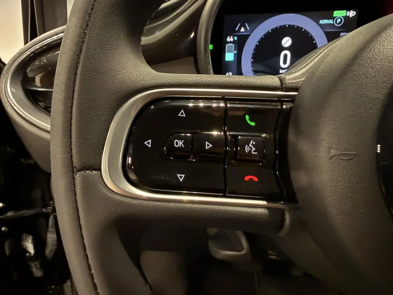 Gros plan sur les commandes gauche du volant noir de la Fiat 500 électrique 2024 avec affichage numérique en arrière-plan.