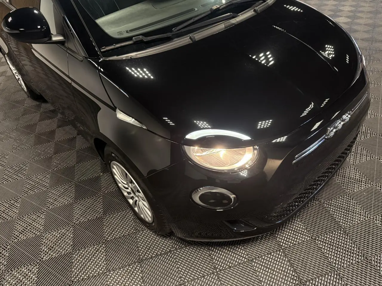 Vue 3/4 avant droit d'une Fiat 500 électrique noire avec phare avant allumé et jantes acier 15 pouces.