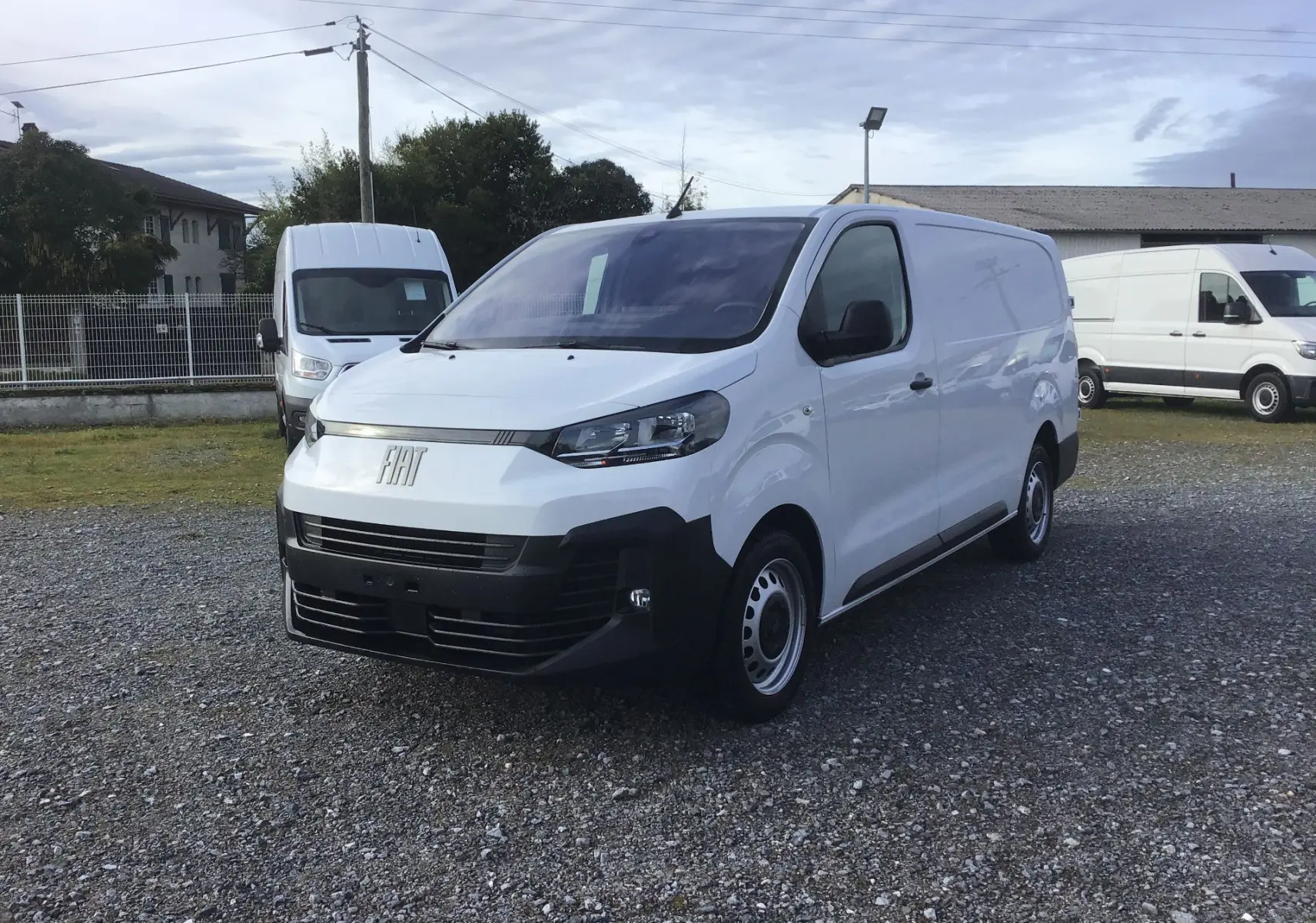 Fourgon FIAT Scudo blanc en 3/4 avant droit, avec pare-chocs noir et jantes acier, stationné sur un sol gravillonné.