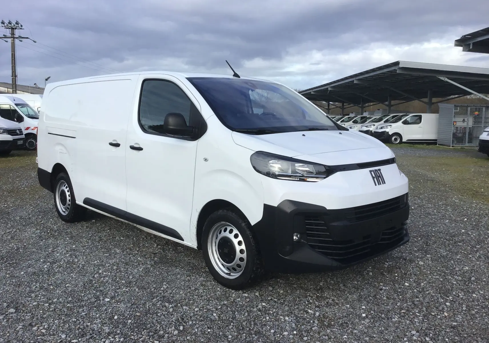 Fourgon FIAT Scudo blanc en 3/4 avant droit, avec pare-chocs noirs et jantes acier, stationné sur sol gravillonné.