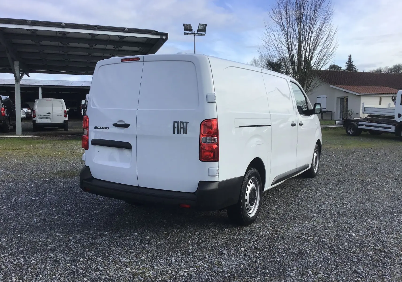 Vue 3/4 arrière droite d’un fourgon FIAT Scudo blanc avec logo FIAT et feux arrière rouges sur un sol gravillonné.