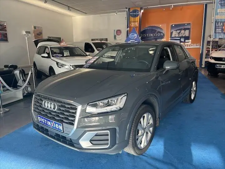 Audi Q2 gris Daytona métal vue 3/4 avant droit dans un showroom avec sol bleu et panneaux publicitaires.
