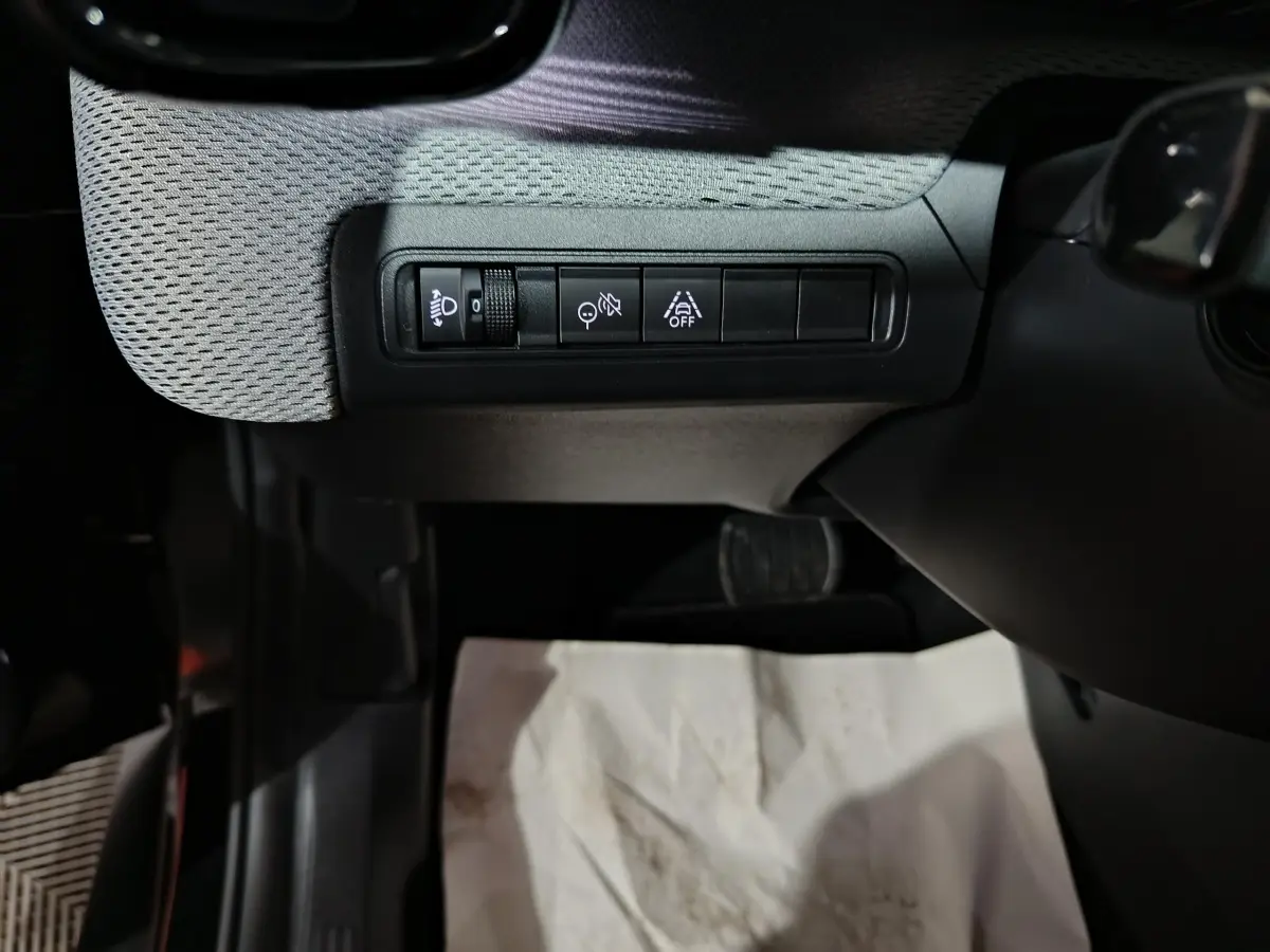 Gros plan sur les commandes d'assistance et réglage des phares côté gauche du tableau de bord d'une Citroën C3 noire hybride 2025.