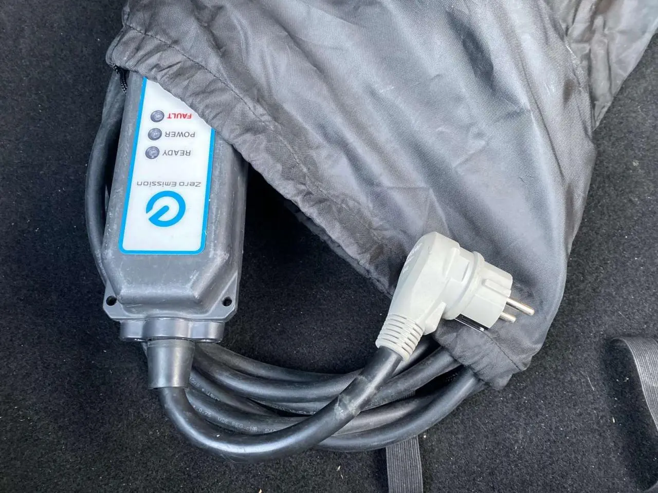 Câble de recharge électrique noir partiellement couvert dans un sac gris, posé sur un tapis de coffre noir.