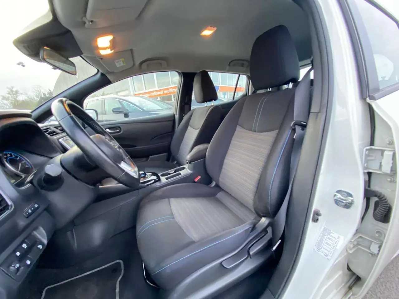 Vue intérieure côté conducteur du Nissan Leaf blanc 2020, sièges tissu noir avec surpiqûres bleues et tableau de bord visible.