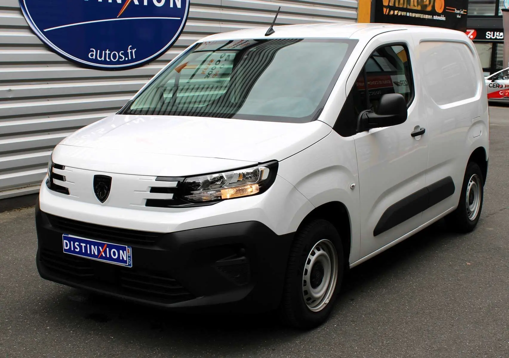 Peugeot Partner Fourgon blanc vue 3/4 avant droit avec calandre noire et feux allumés sur parking.