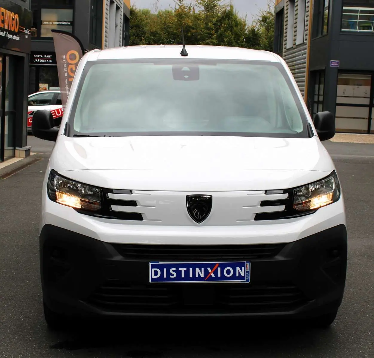 Vue de face du Peugeot Partner Fourgon blanc 2025 avec feux allumés et logo noir distinctif sur la calandre.