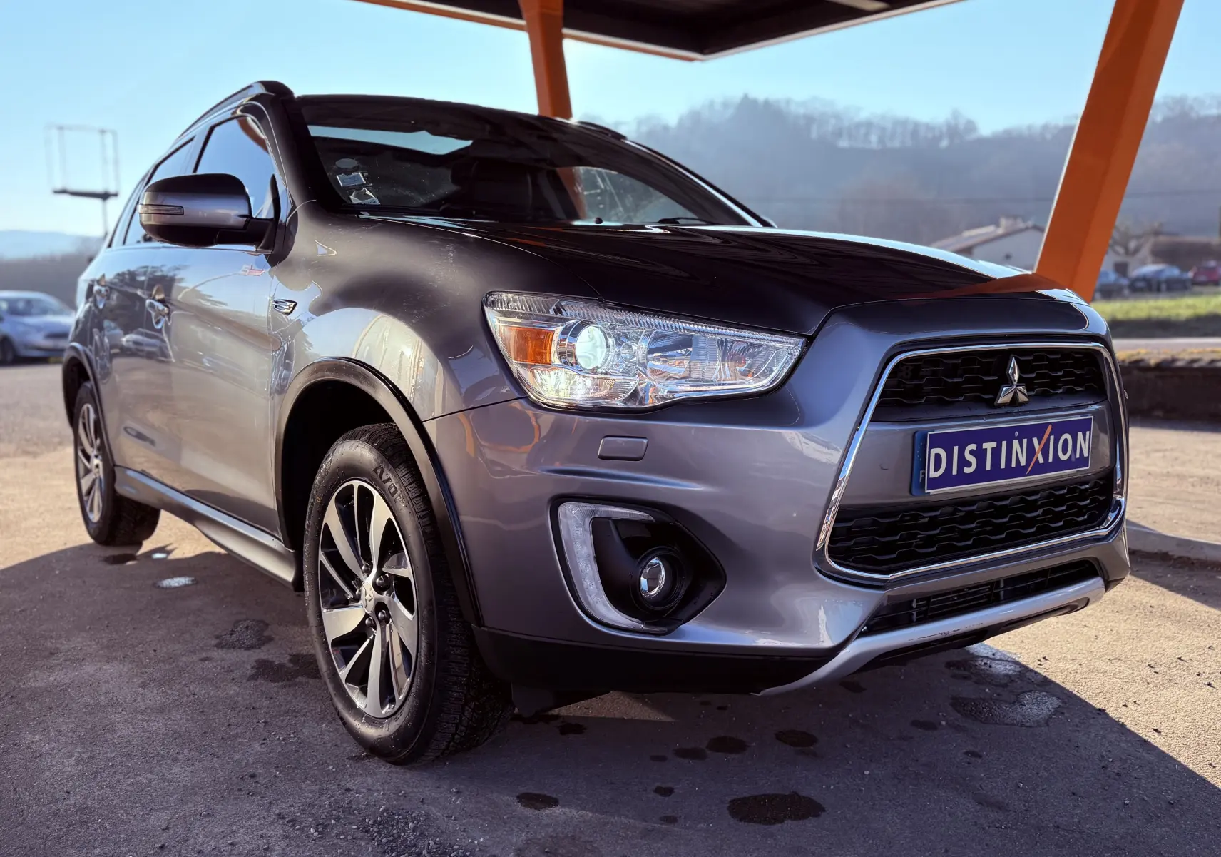 Mitsubishi ASX gris en 3/4 avant droit, avec calandre noire et phares allumés sous un abri.