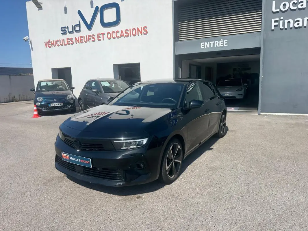 Opel Astra noire 2024 vue 3/4 avant droit, avec jantes alliage 17 pouces et vitres surteintées, stationnée devant un garage.