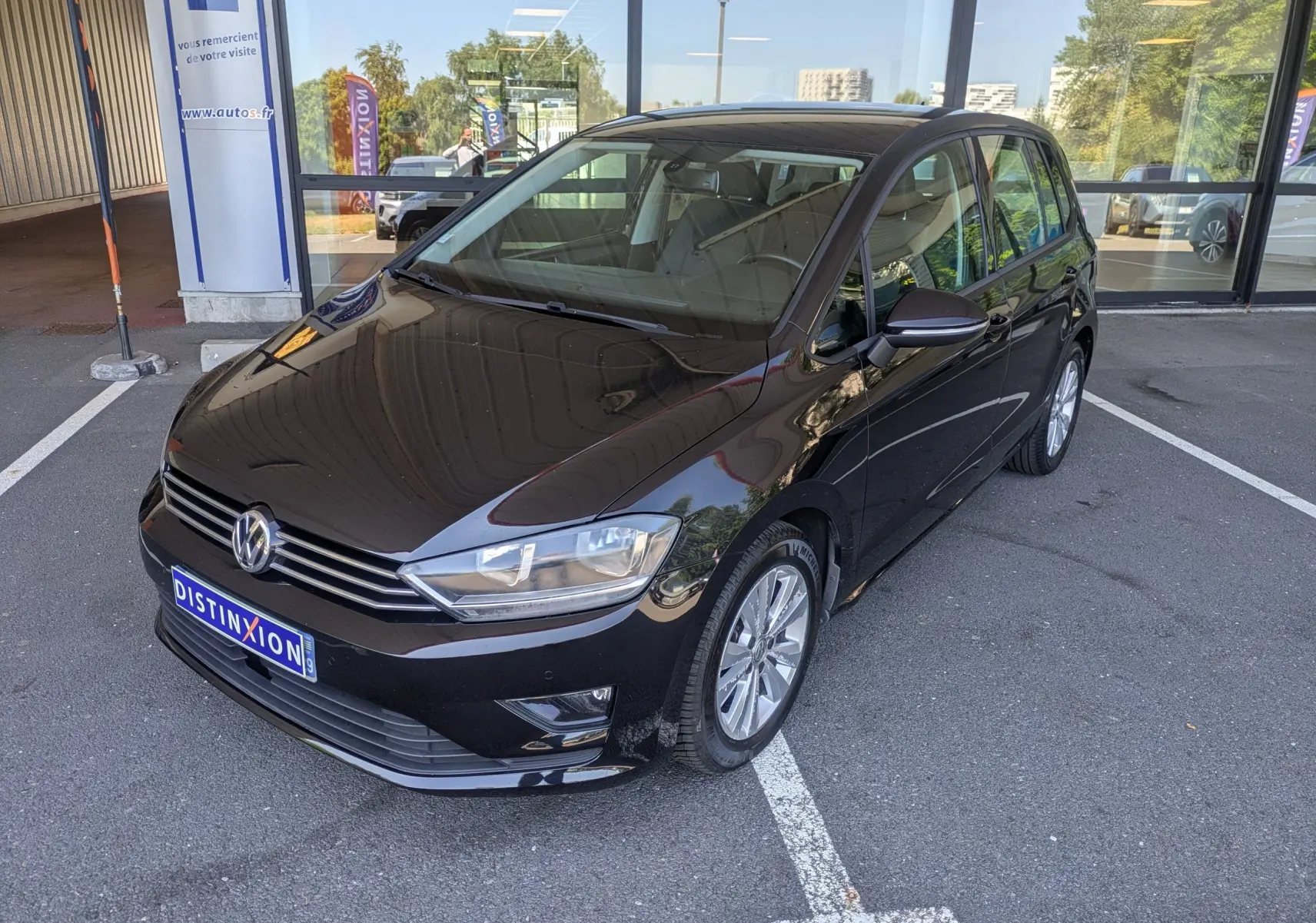Volkswagen Golf Sportsvan noir vue 3/4 avant droit, garée devant une vitrine avec plaques Distinxion.