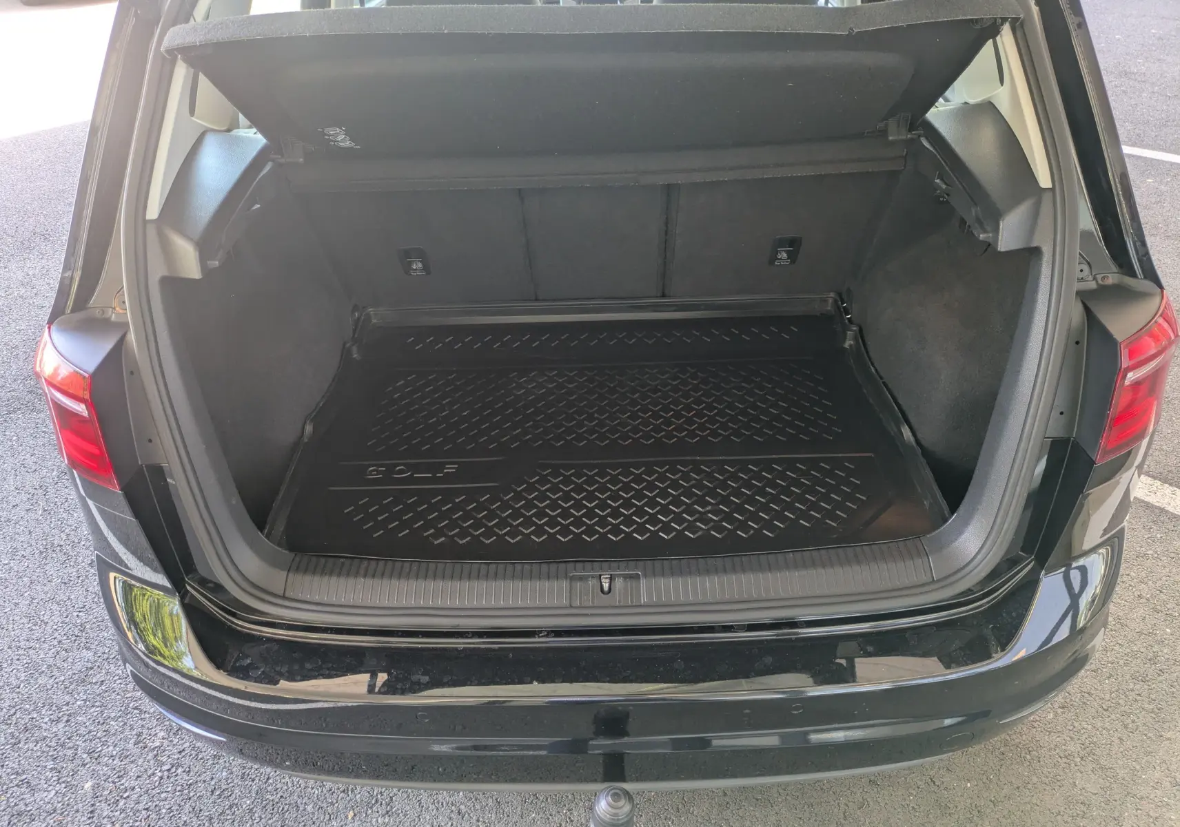 Coffre ouvert vu de l'arrière d'une Volkswagen Golf Sportsvan noire, avec tapis de coffre marqué "GOLF".