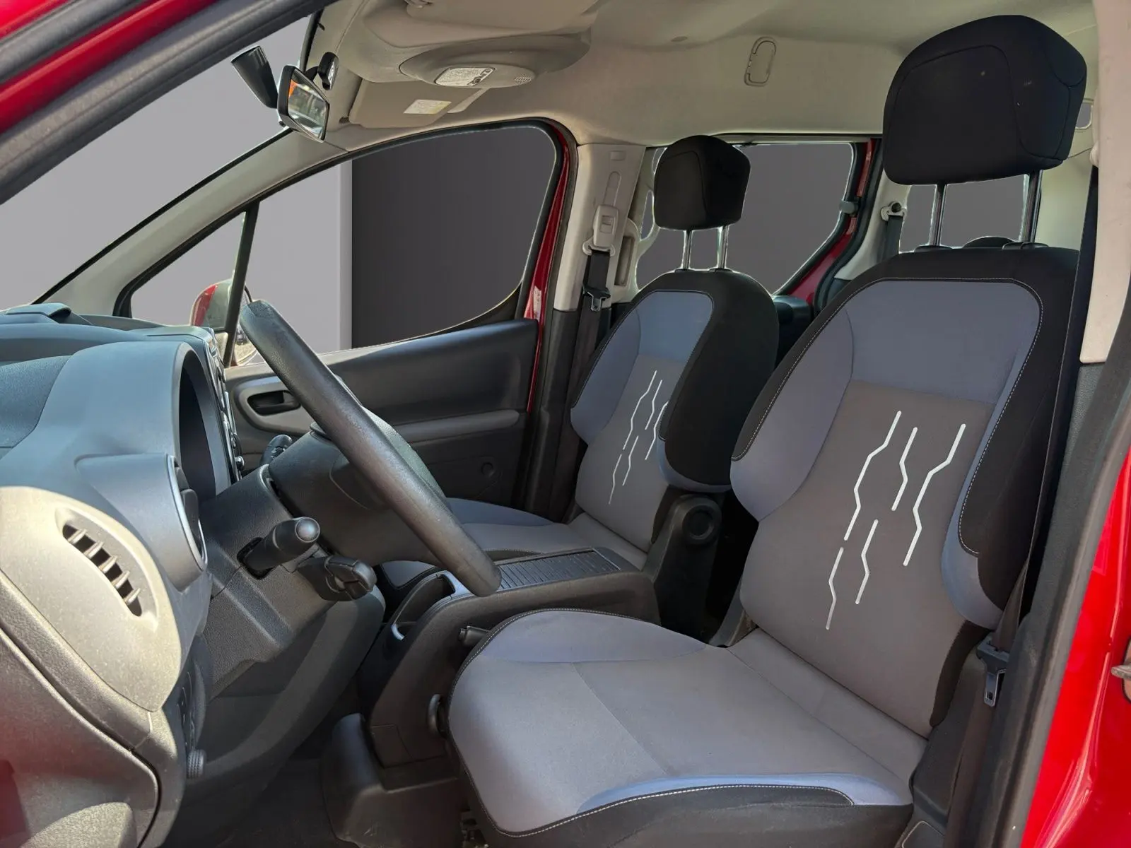 Vue intérieure côté conducteur du Peugeot Partner Tepee rouge 2016, sièges avant gris avec motifs blancs et volant réglable.