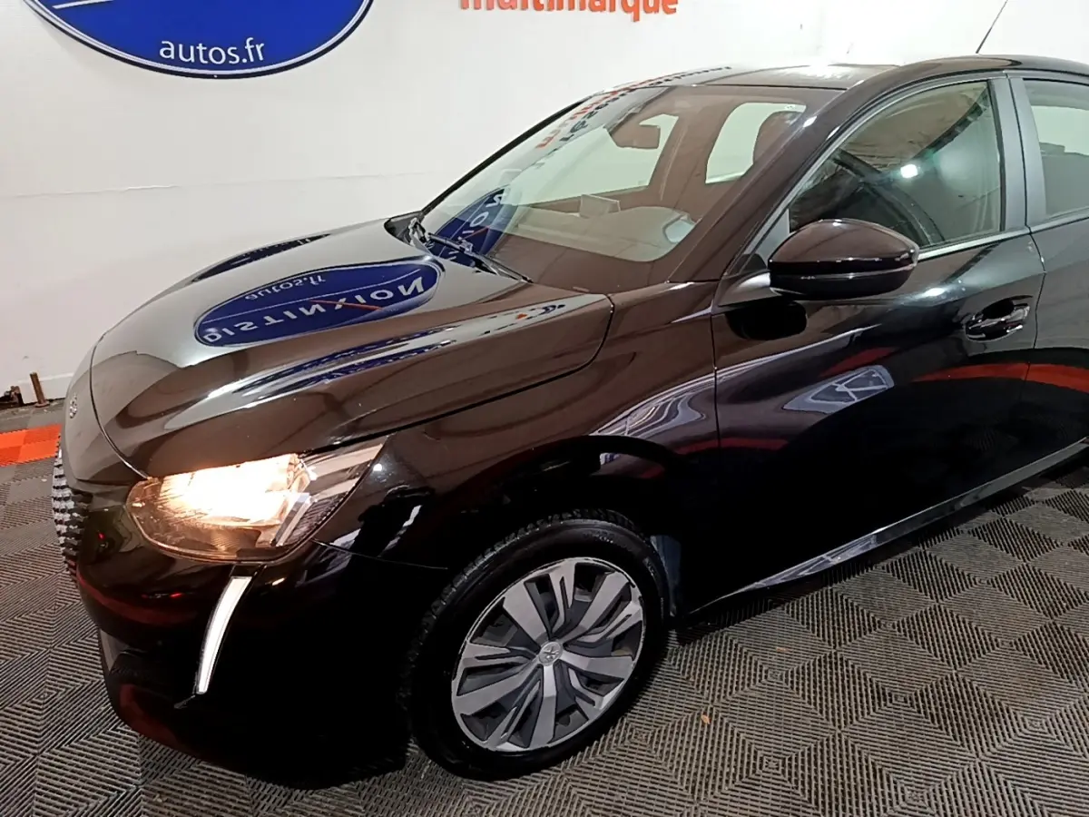 Vue 3/4 avant droit d'une Peugeot 208 noire avec phares allumés et jantes distinctives en intérieur showroom.