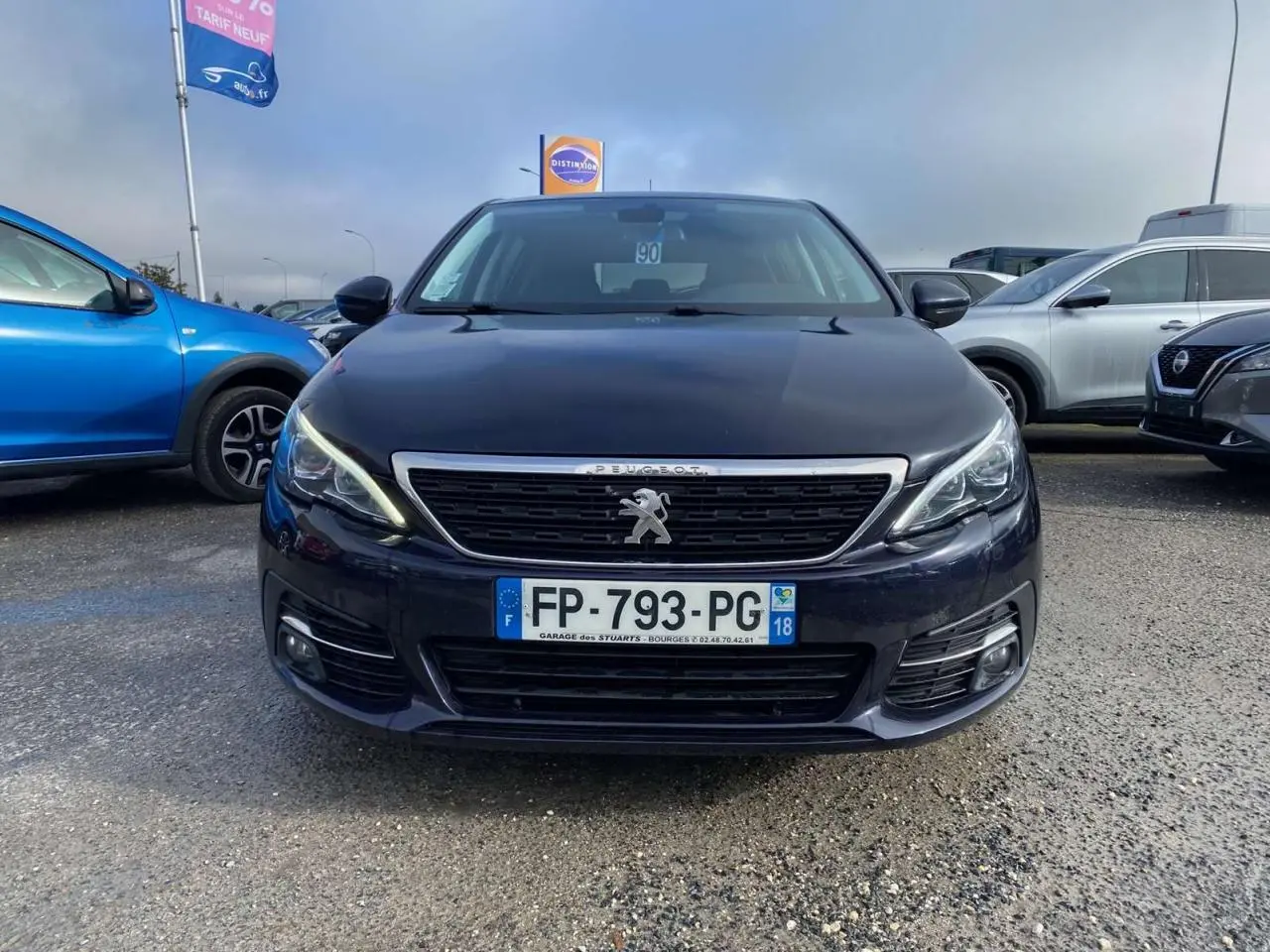 Vue frontale d'une Peugeot 308 bleu foncé avec calandre chromée et feux LED allumés sur un parking extérieur.