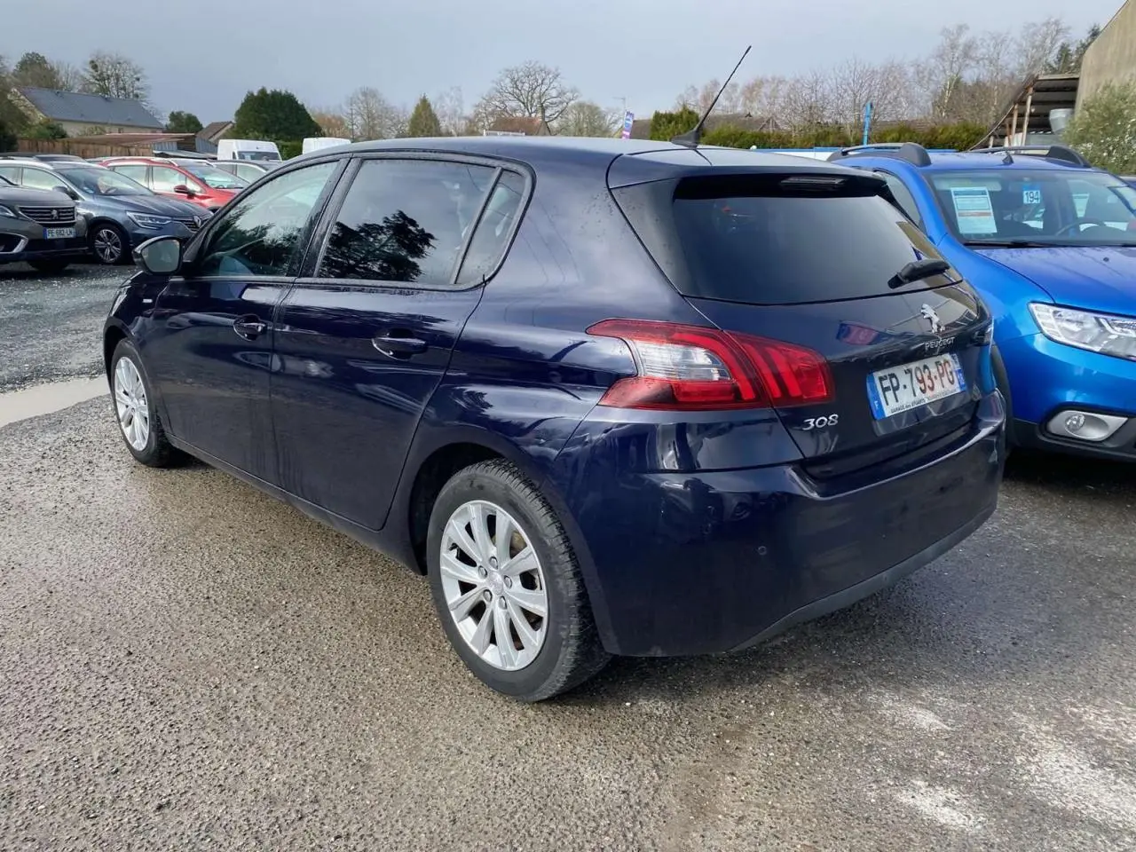 Peugeot 308 bleu foncé vue 3/4 arrière droit, avec feux arrière et jantes visibles sur parking extérieur.