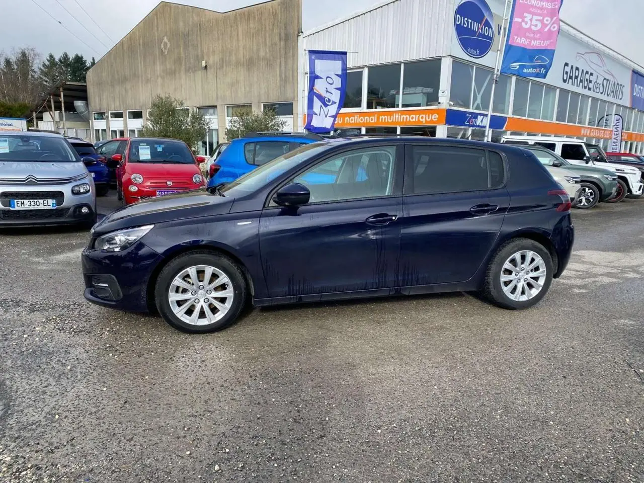 Vue de profil côté gauche d'une Peugeot 308 2018 bleu foncé métallisé, stationnée devant un garage multimarque.