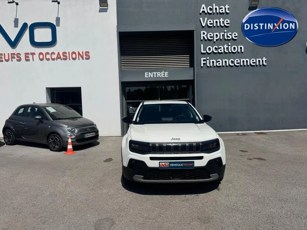 Vue avant d'une Jeep Avenger électrique blanche stationnée devant un bâtiment commercial avec enseignes visibles.