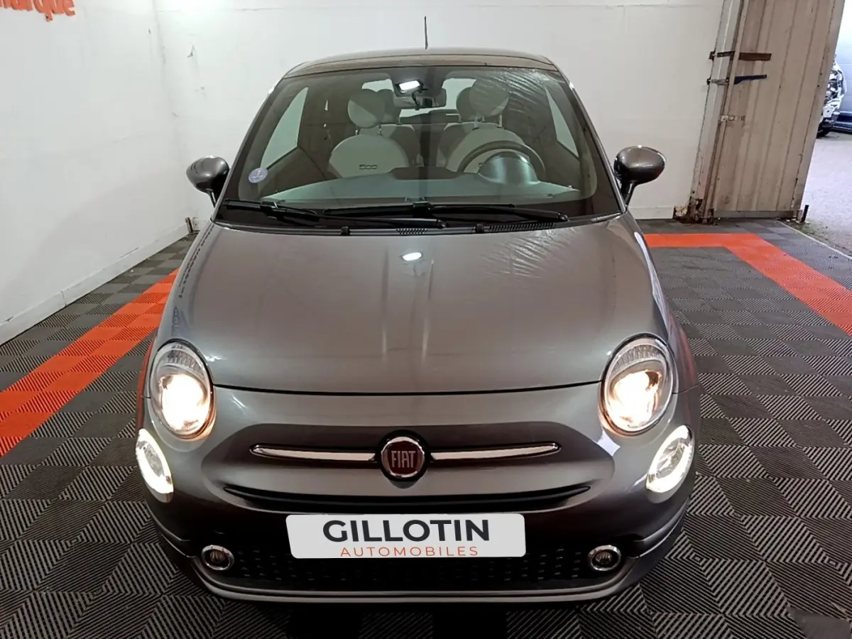 Vue frontale d'une Fiat 500 gris foncé avec phares allumés dans un garage intérieur.