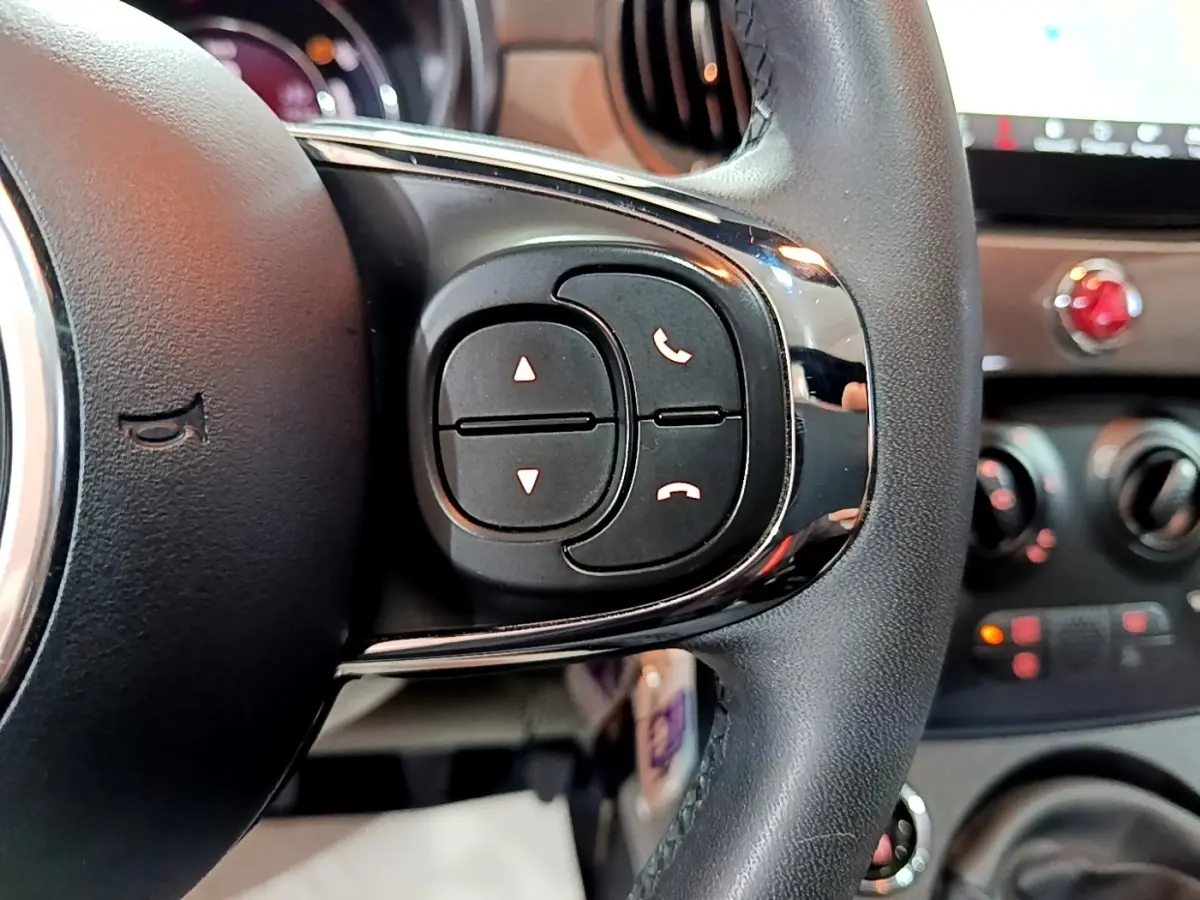 Gros plan sur les commandes du volant cuir noir de la Fiat 500 Dolcevita 2022, avec tableau de bord flou en arrière-plan.