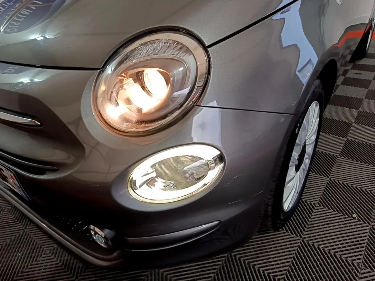 Gros plan sur l'avant droit d'une Fiat 500 gris foncé avec phare principal allumé et jante blanche visible.