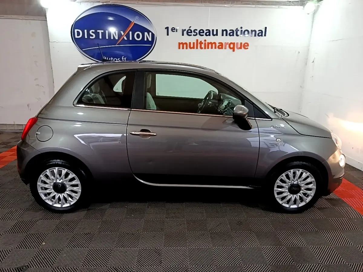 Profil droit d'une FIAT 500 gris foncé 2022 avec jantes alliage et intérieur clair visible en arrière-plan showroom.