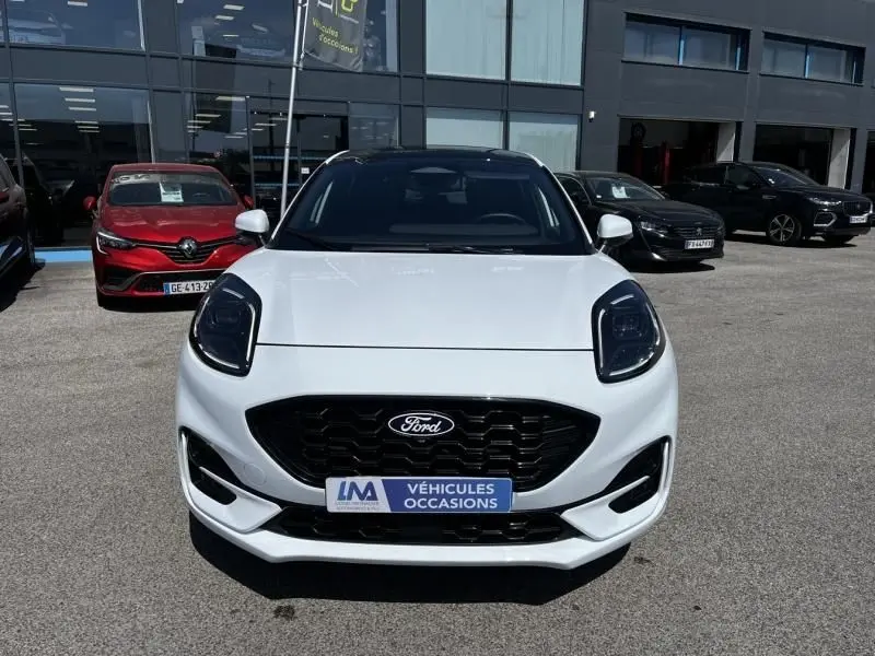 Vue frontale d'un Ford Puma blanc 2025 avec calandre noire et badge Ford, stationné devant un bâtiment commercial.