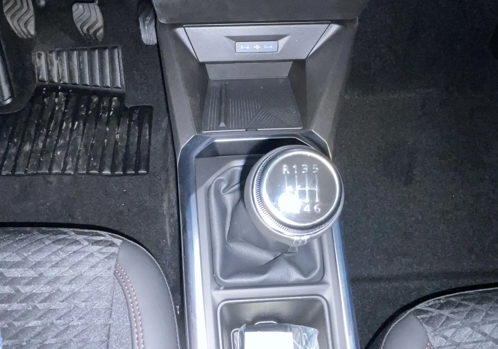 Vue plongeante sur la boîte manuelle et le levier de vitesse de la Renault Clio gris Rafale, avec pédales et sièges partiellement visibles.