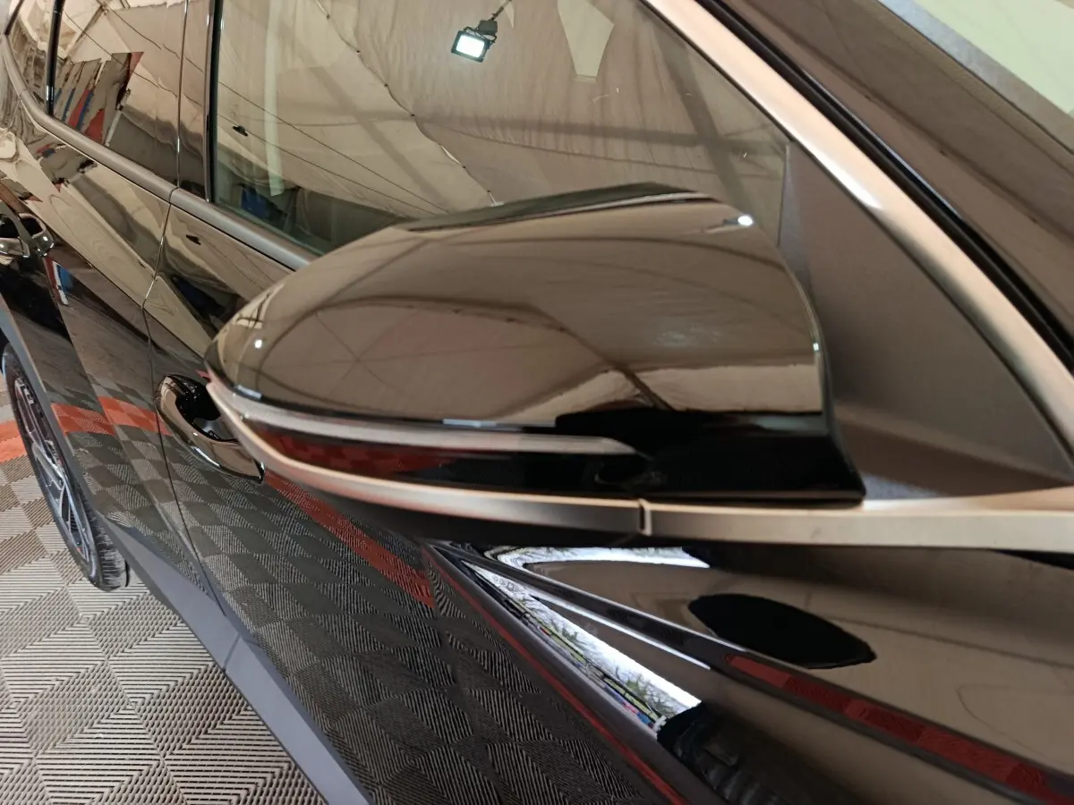Gros plan sur le rétroviseur côté gauche noir brillant du Hyundai Tucson 1.6 Hybrid 2025, avec reflet de l’environnement.