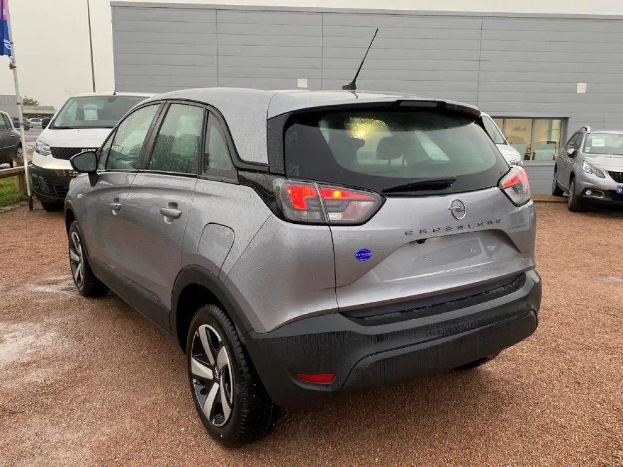 Opel Crossland gris métallisé vu en 3/4 arrière gauche sous la pluie, avec feux arrière allumés et jantes bicolores.