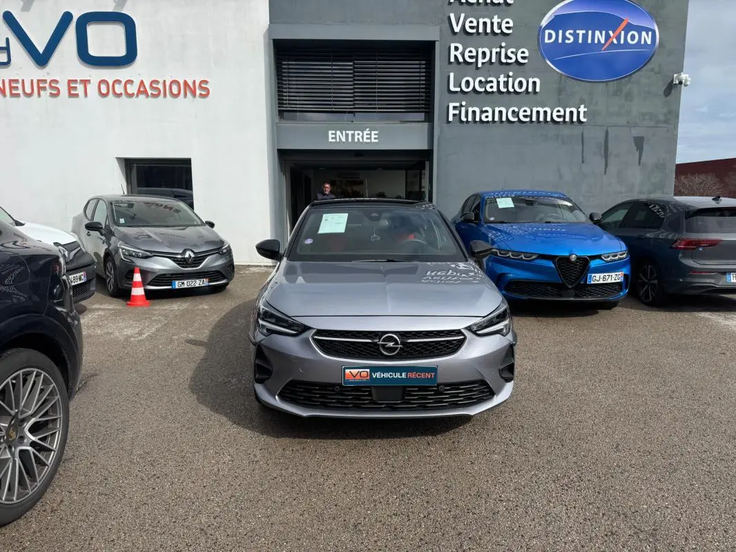 Vue avant d'une Opel Corsa gris clair 2022 garée devant un concessionnaire avec logo et panneaux visibles.