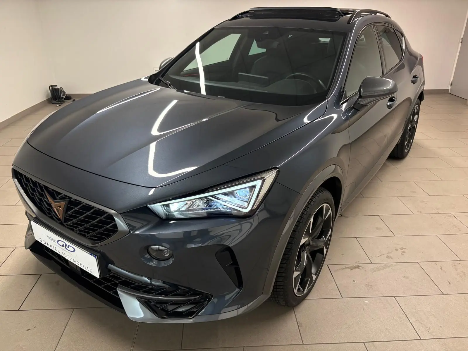 SUV CUPRA Formentor gris magnétique vu en 3/4 avant droit, avec phares allumés et jantes noires distinctives.