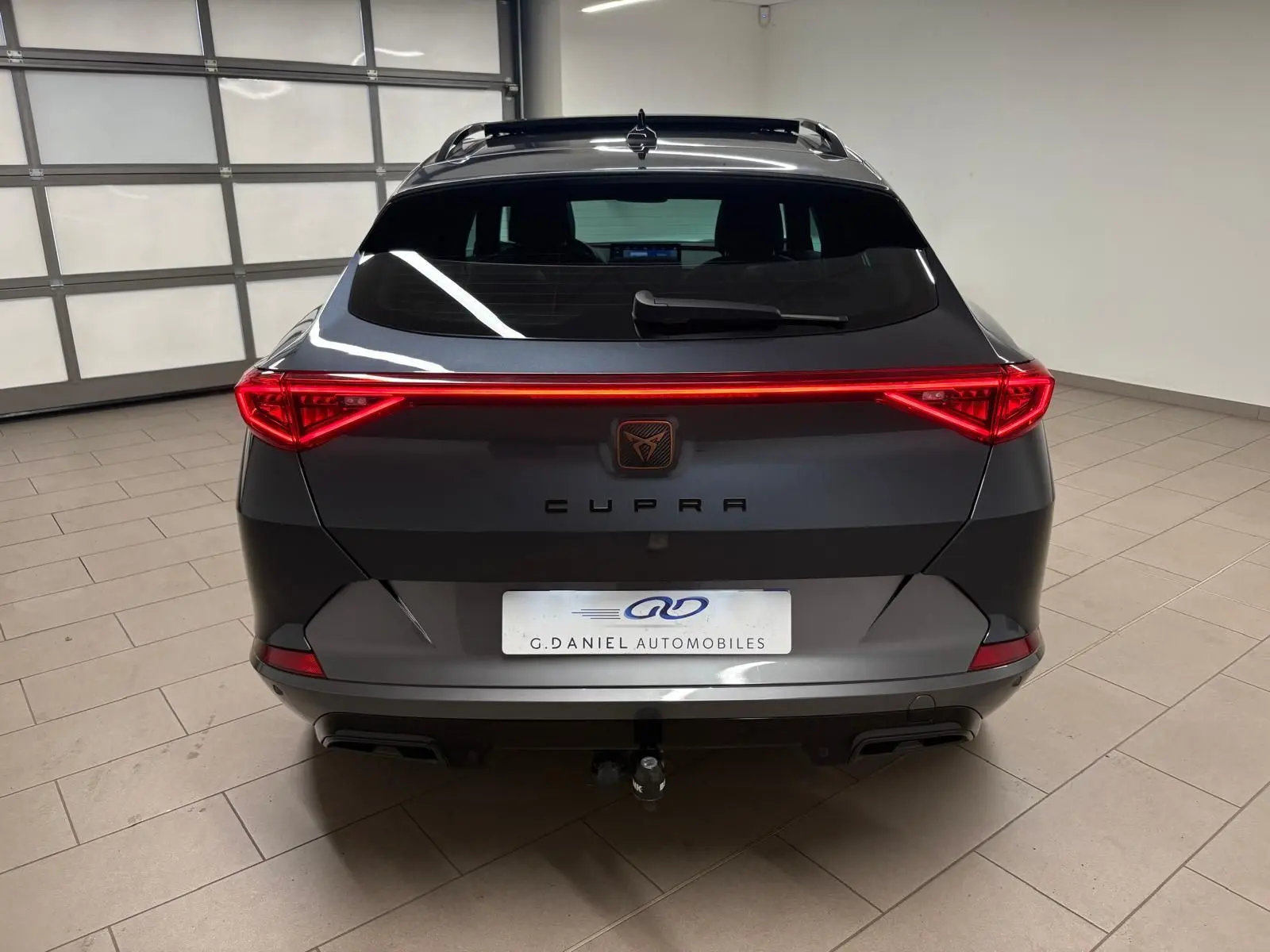 Vue arrière d'un CUPRA Formentor gris magnétique 2023 avec feux LED reliés et attelage visible en intérieur showroom.