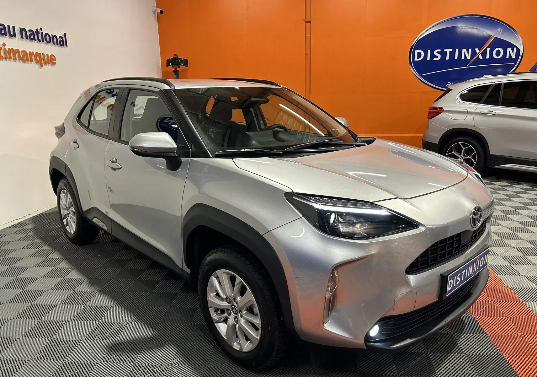 Toyota Yaris Cross gris minéral vue 3/4 avant droit en intérieur avec phares et feux de jour allumés.