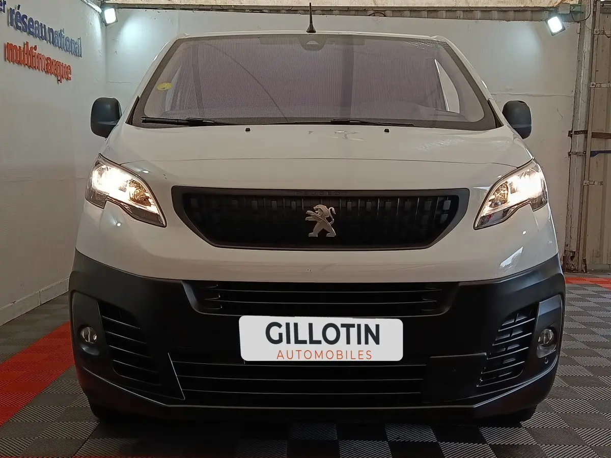 Vue de face d'un Peugeot Expert Fourgon blanc avec calandre noire et phares allumés dans un garage.