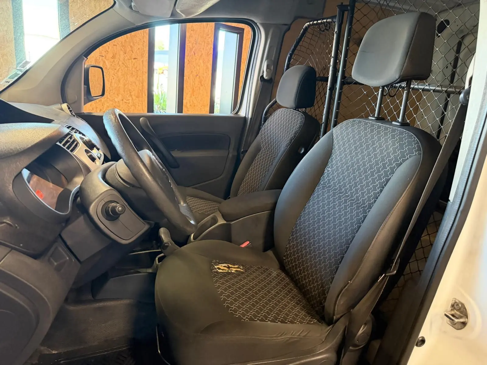 Vue intérieure côté conducteur du Renault Kangoo blanc 2011, montrant les sièges en tissu noir et la cloison métallique séparatrice.