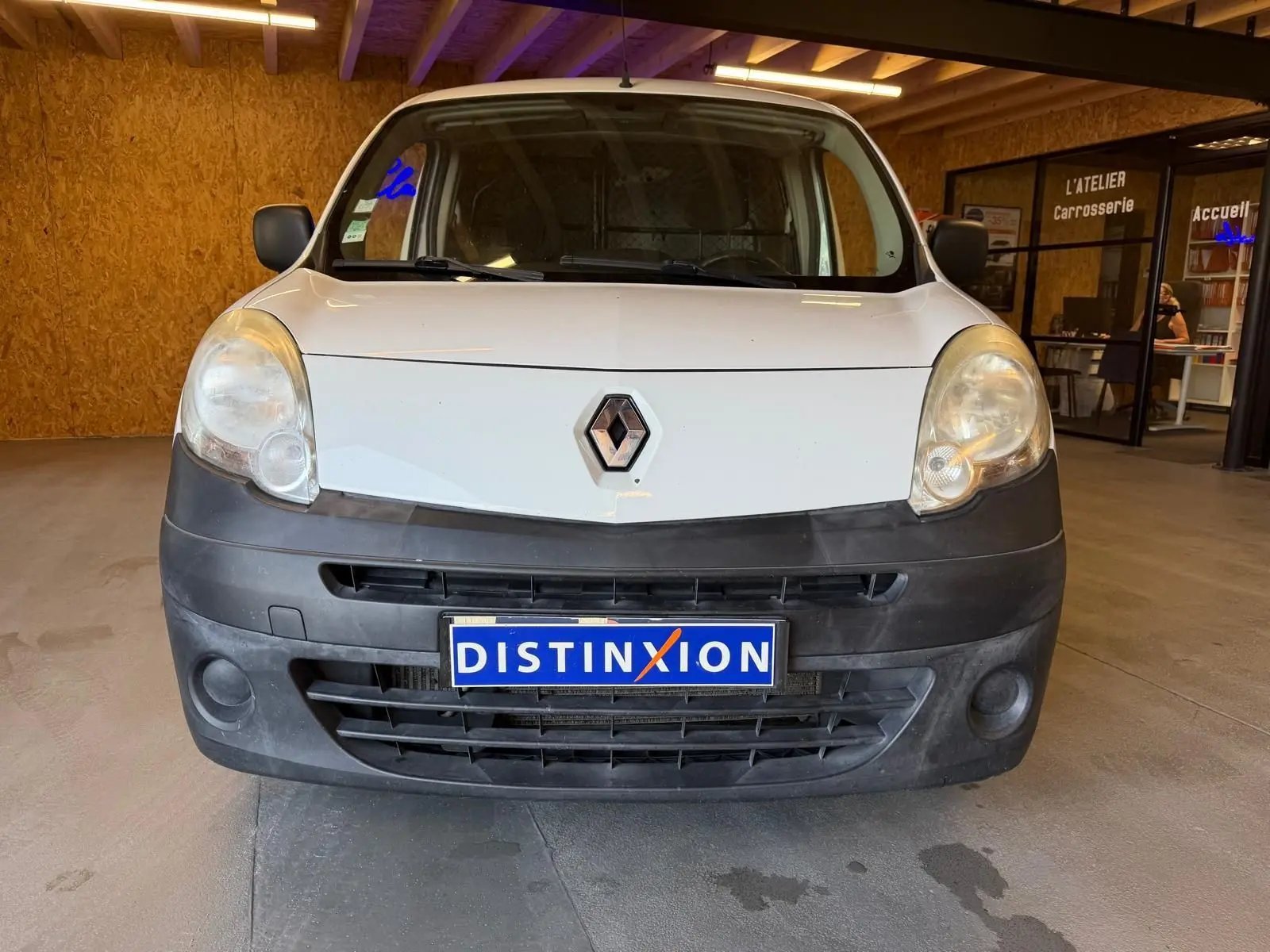 Vue frontale d’un Renault Kangoo blanc 2011 avec pare-chocs noir mat et plaque Distinxion en intérieur d’atelier.