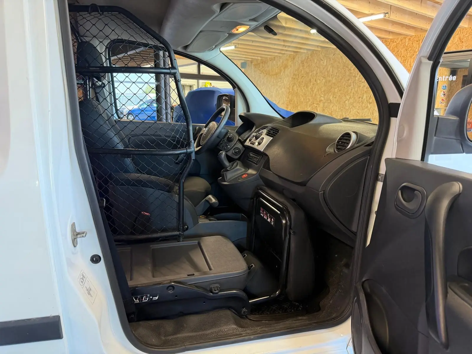 Vue intérieure côté conducteur du Renault Kangoo blanc 2011 avec cloison métallique séparant l'habitacle et siège en tissu noir.