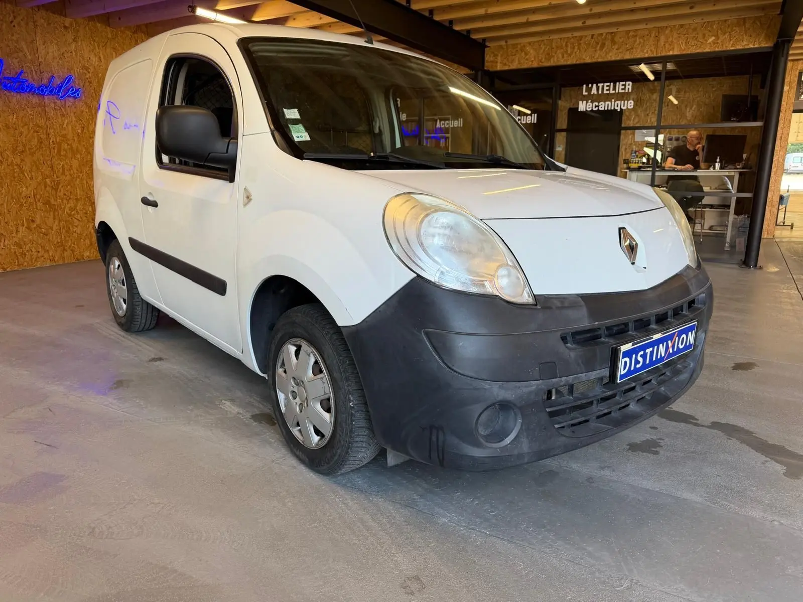 Vue 3/4 avant gauche d’un Renault Kangoo blanc de 2011 avec pare-chocs noir et plaque Distinxion en intérieur d’atelier.